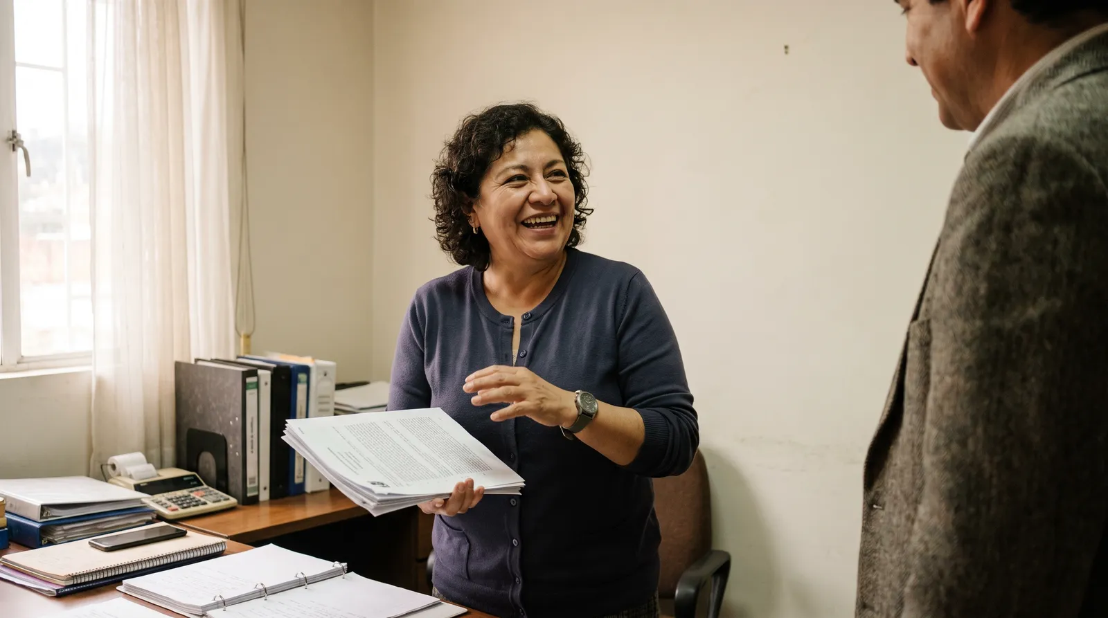 A woman laughs while handing a stapled document to a man standing in her office.