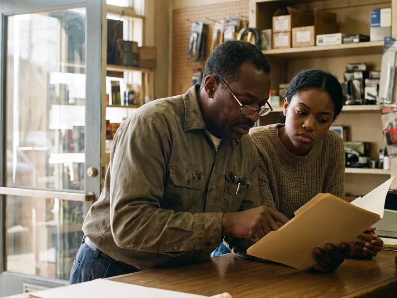 An older man and a young woman lean over an open folder together behind a shop counter.