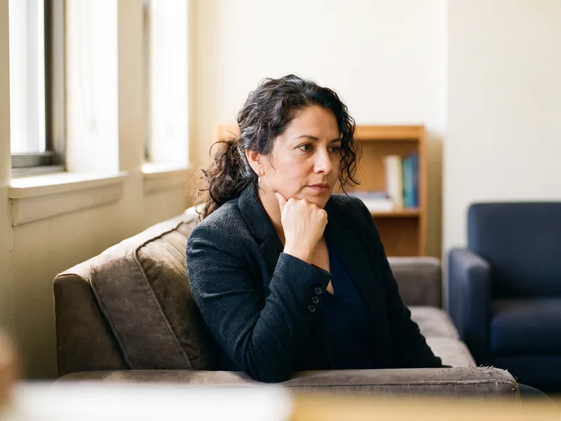 A woman in a blazer sits on a sofa listening to someone across from her, hand at her chin.