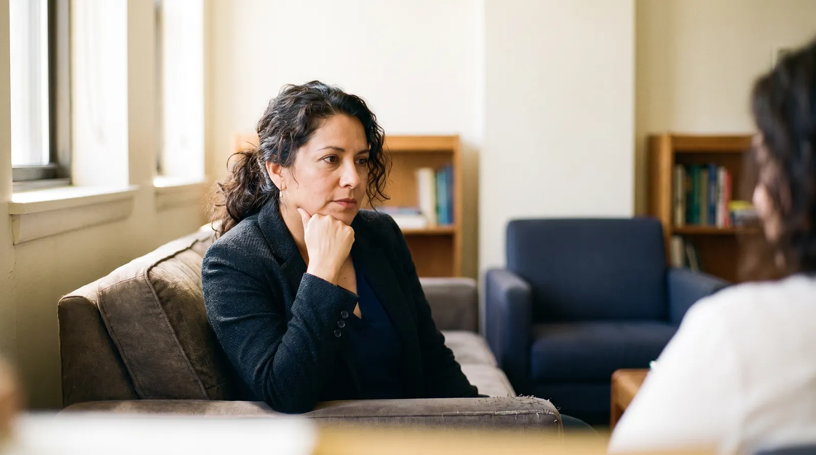 A woman in a blazer sits on a sofa listening to someone across from her, hand at her chin.